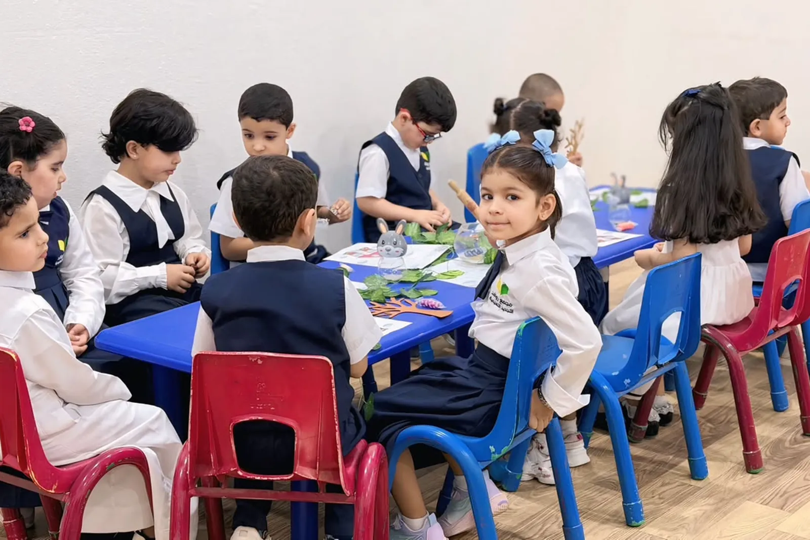 أطفال الروضة حول طاولة نشاط في أحد فصول المجمع، بزيّ المجمع وشعاره